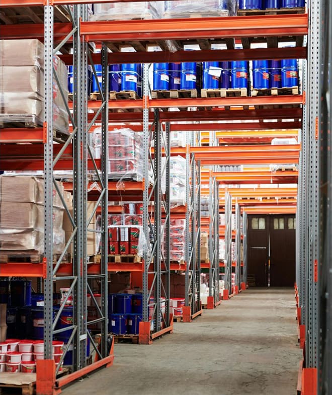 Instalación de almacenamiento de productos químicos con estanterías altas de acero que contienen paletas de cajas, contenedores y barriles, organizados a lo largo de amplios pasillos de hormigón.